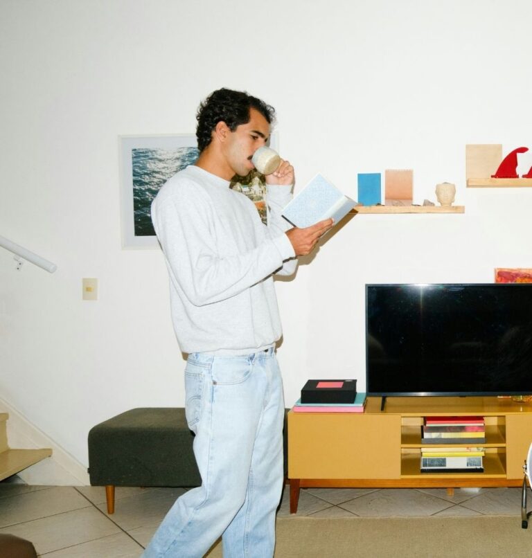 young guy reading a book while walking through house drinking coffee
