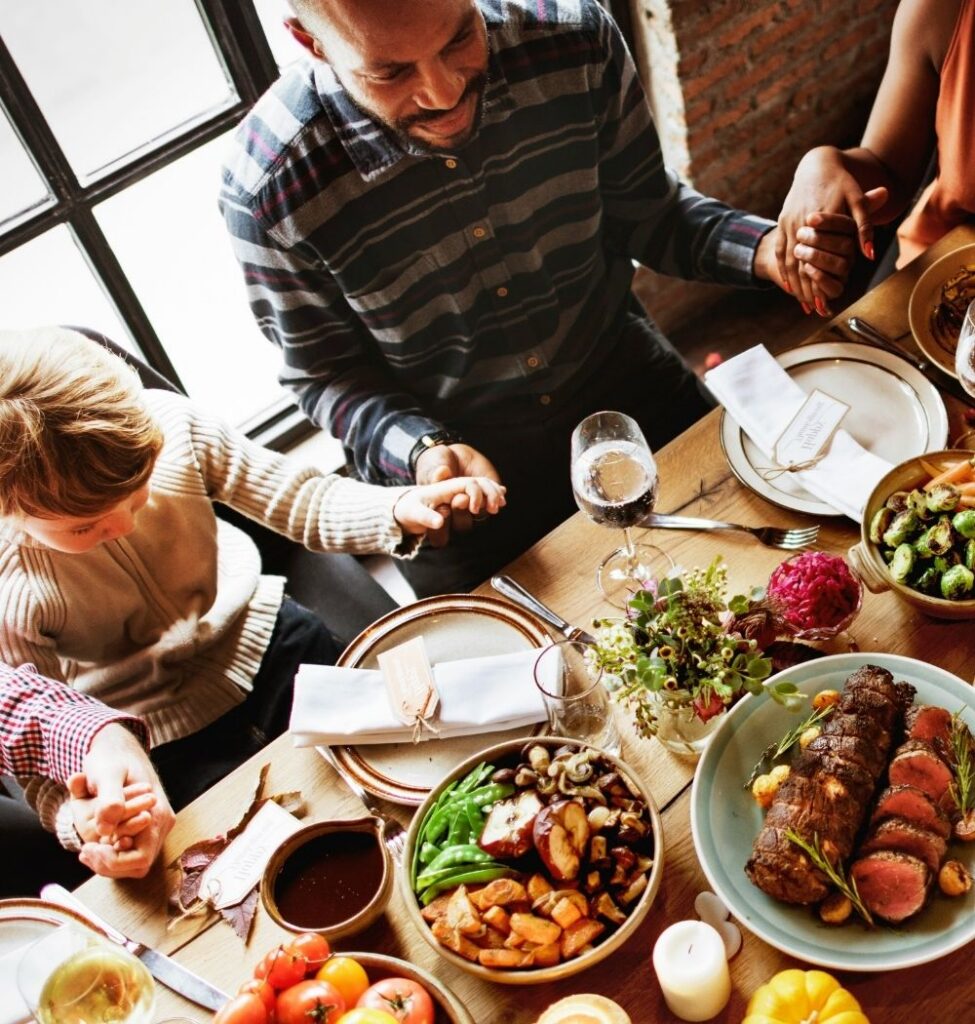 family around table full of food saying grace or prayer while holding hands