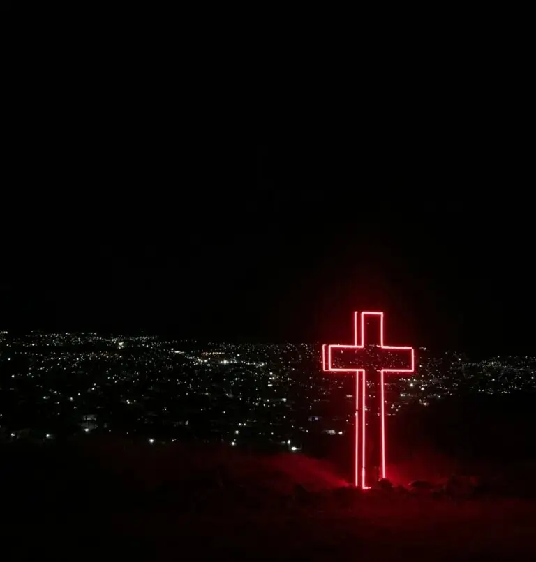 night sky with a red neon cross sculpture in the foreground and lights from a city in the background