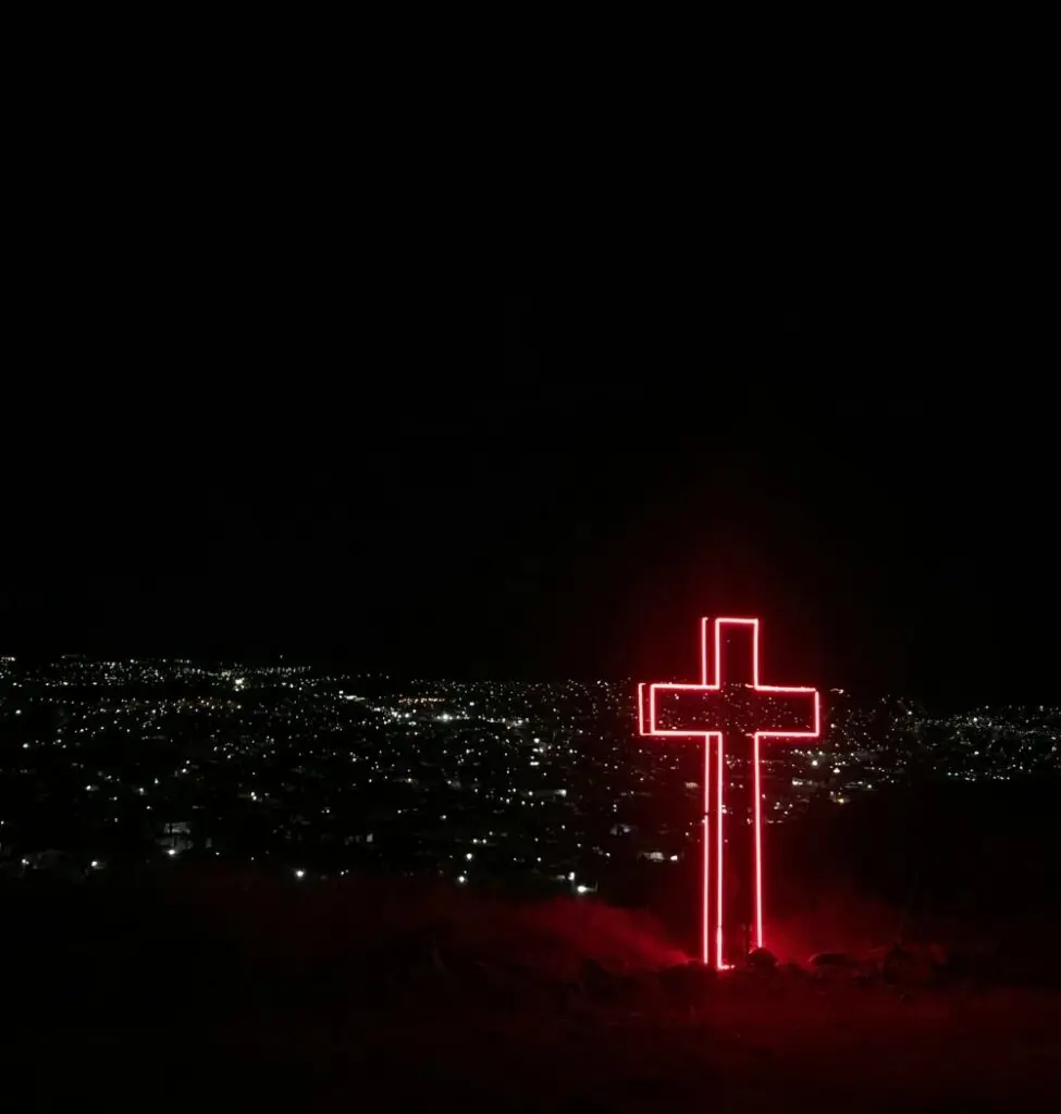 night sky with a red neon cross sculpture in the foreground and lights from a city in the background