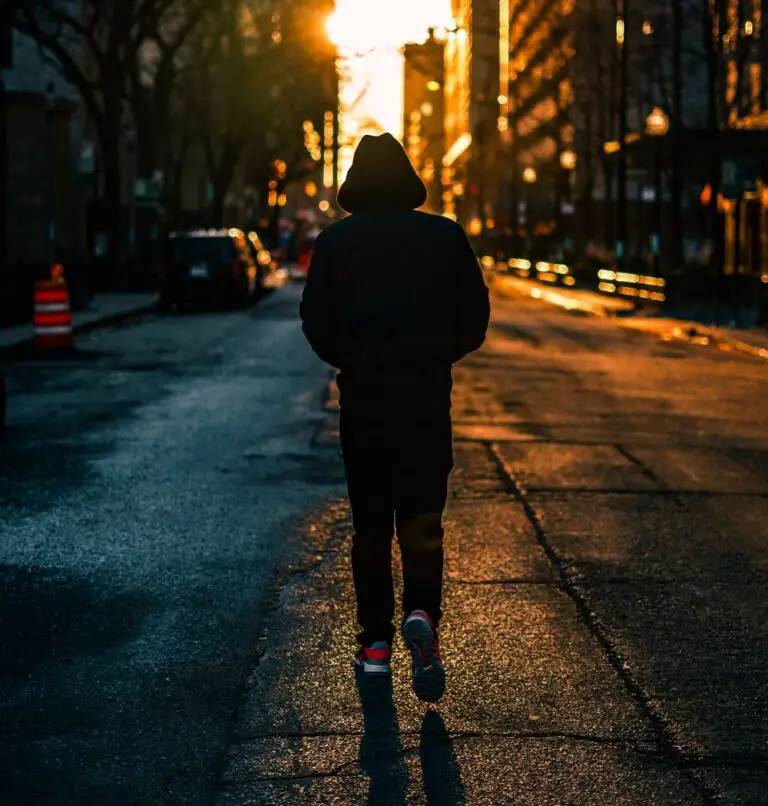 Young Christian man in hoodie walking down a dark city street toward the sunrise.