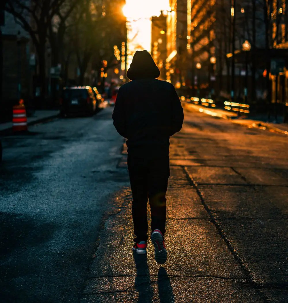 Young Christian man in hoodie walking down a dark city street toward the sunrise.