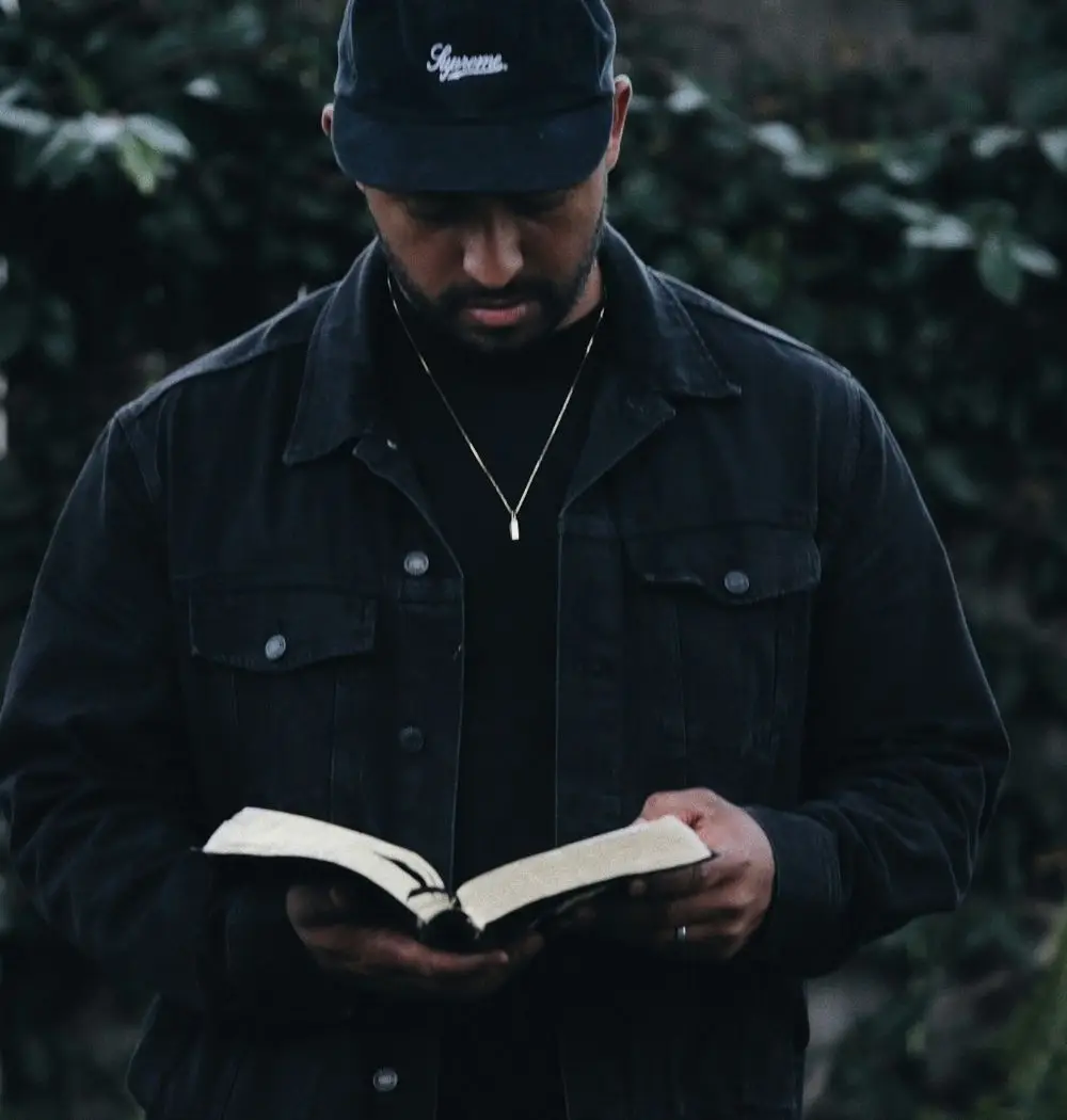 African-American man dressed in black reads Bible while standing against a dark background