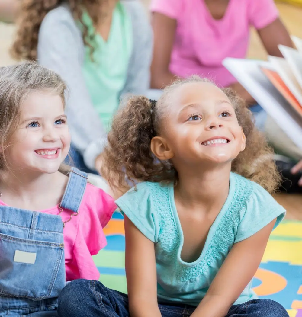 Two young girls in first grade smile early during story time.