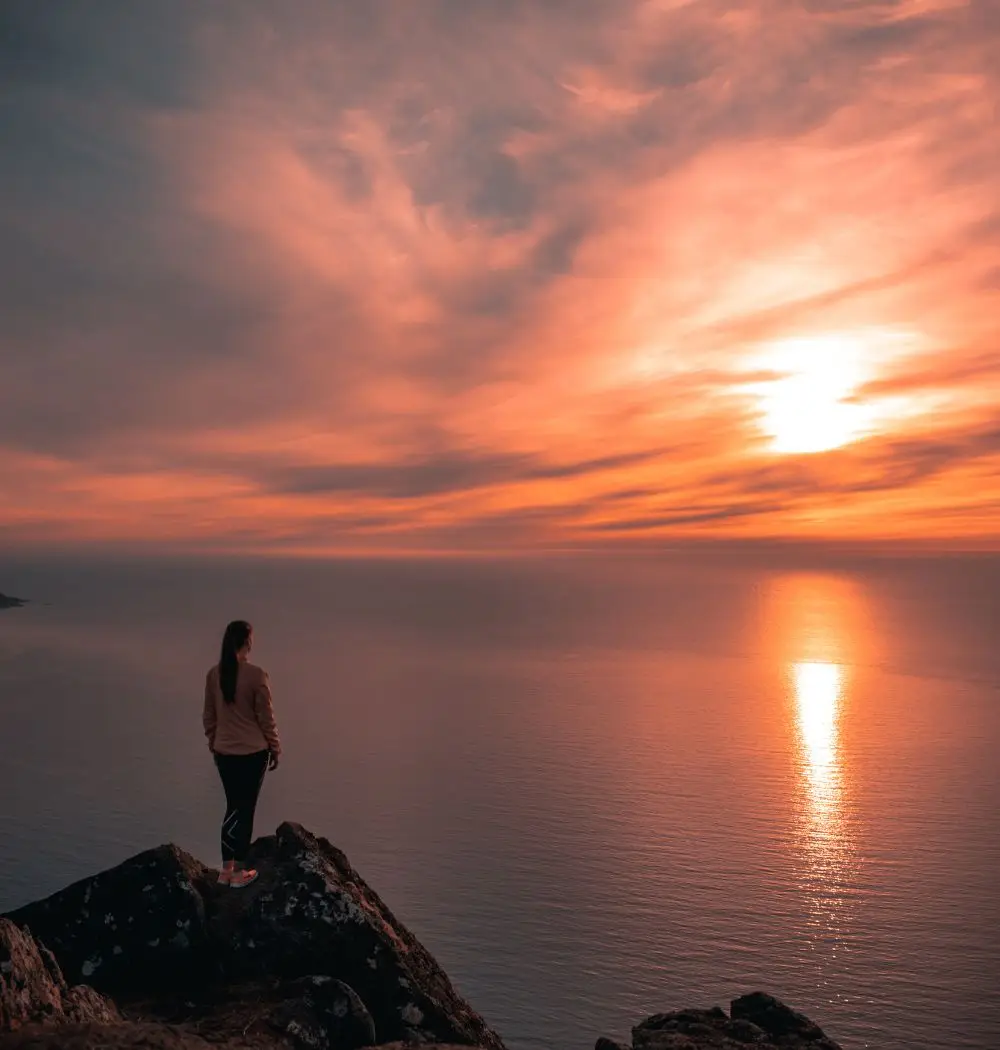 Christian curious woman standing on seaside cliff looking at the sunset